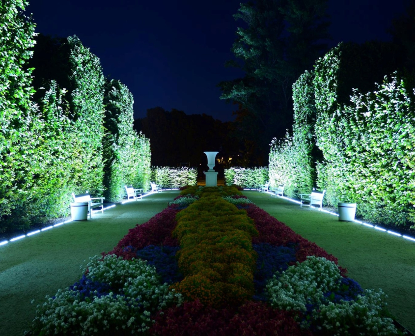 Nighttime garden view with vibrant flower beds leading to a white vase, flanked by meticulously trimmed hedges illuminated by commercial landscape lighting. Benches line the path, inviting a peaceful stroll.