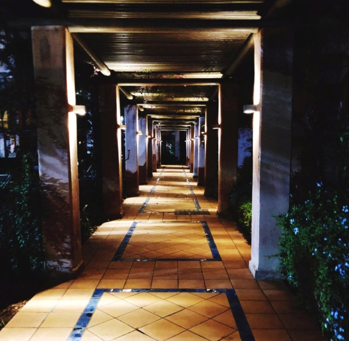 Illuminated walkway with tiled floor and columns under a pergola at night. The commercial landscape lighting creates a warm, inviting atmosphere.