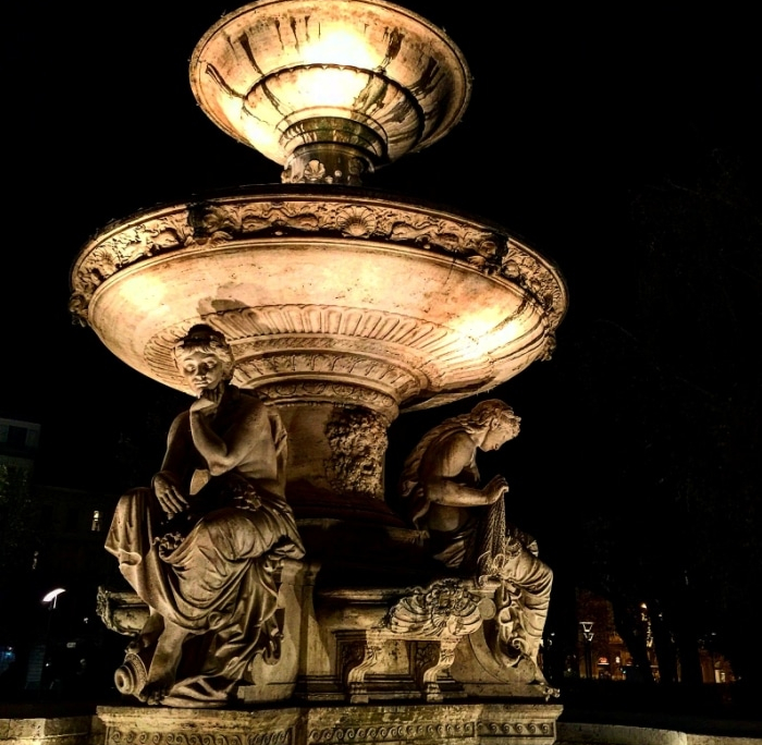 Illuminated stone fountain at night, featuring sculpted figures. Professional landscape lighting highlights the intricate details, creating a dramatic effect. Regular pressure washing can help maintain its beauty.