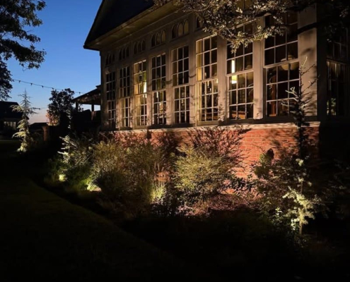 Exterior view of a home at dusk featuring landscape lighting installation. Warm lights illuminate the bushes along the foundation, highlighting the architecture and creating a welcoming ambiance.