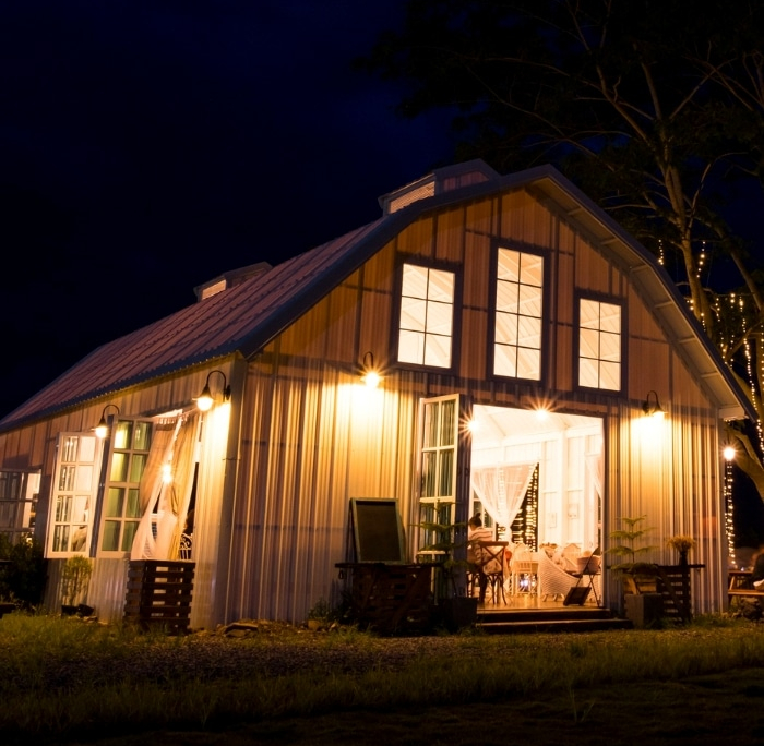Evening view of a barn-style home illuminated by landscape lighting installation, showcasing warm light spilling from large windows and string lights in the surrounding trees.
