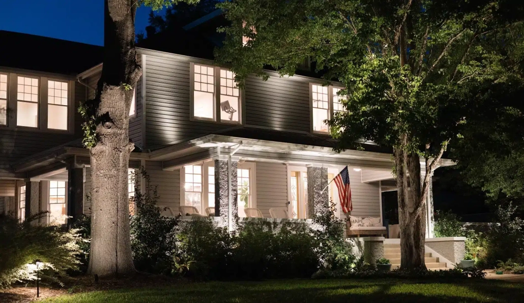 Suburban home at twilight with warm lighting highlighting the facade and surrounding trees. An American flag hangs on the porch, accentuating the home's welcoming feel.