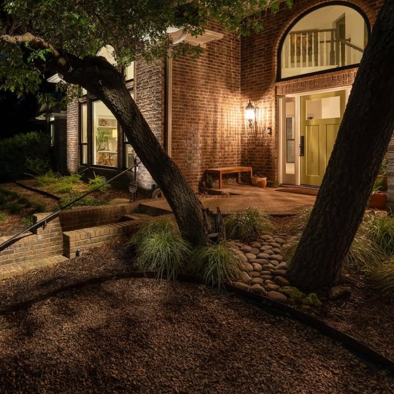 Exterior of a brick home at night, showcasing outdoor lighting design. A warm glow illuminates the front door and a bench, highlighting the landscaping and architectural details.