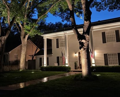 Elegant white home at dusk showcases professional outdoor lighting design, highlighting architectural details and mature trees. American flag displayed on the porch.