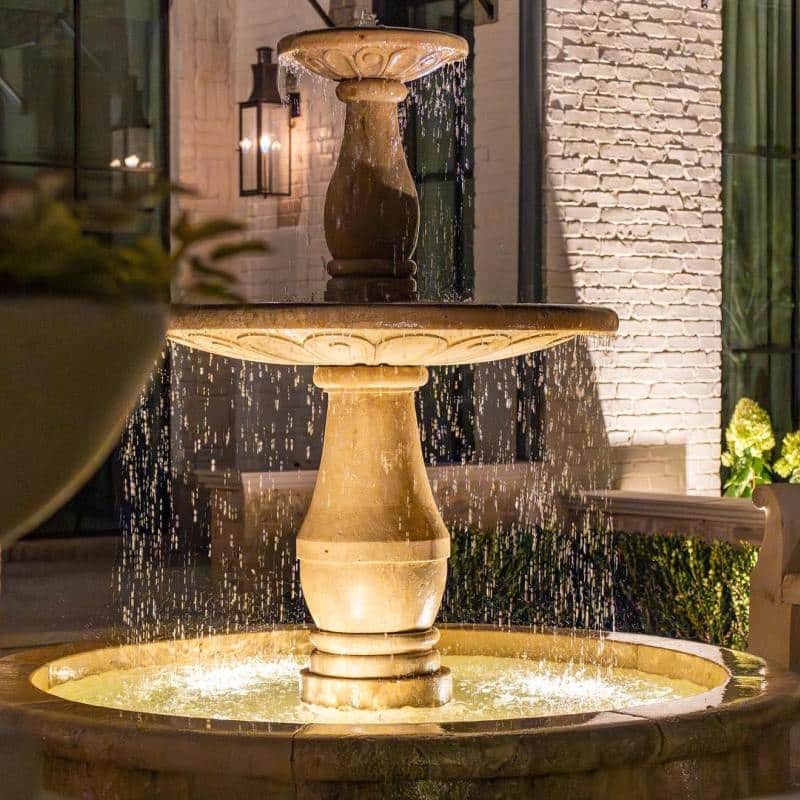 Illuminated tiered stone fountain provides a focal point for outdoor lighting design, with water cascading into a circular basin, set against a brick facade and dark windows.