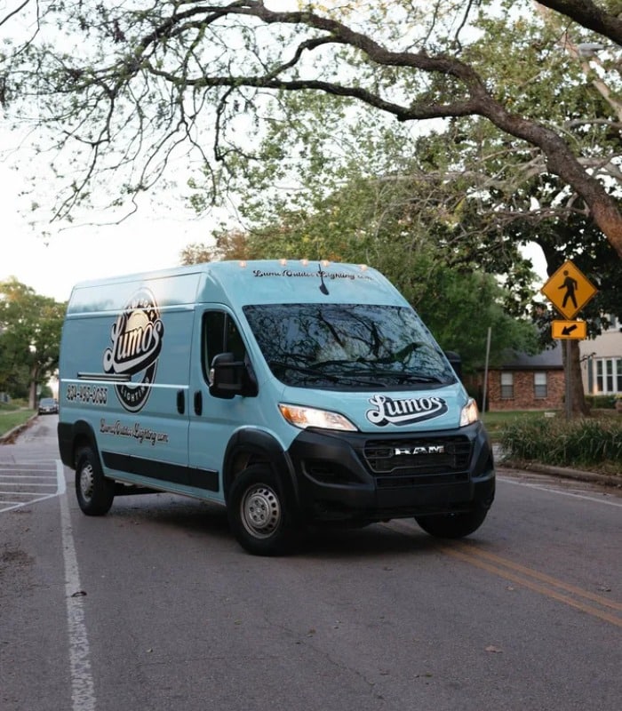 A light blue Lumos Lighting RAM van drives down a tree-lined street, promoting outdoor lighting maintenance services. The van prominently displays the company logo and contact information.