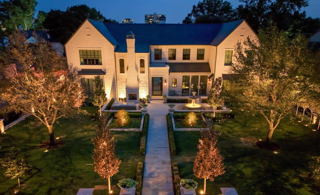 Elegant white brick home illuminated by Lumos Lighting at dusk. The manicured lawn features a symmetrical garden path, mature trees, and a central fountain, all beautifully lit.