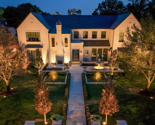 Elegant white brick home illuminated by Lumos Lighting at dusk. The manicured lawn features a symmetrical garden path, mature trees, and a central fountain, all beautifully lit.