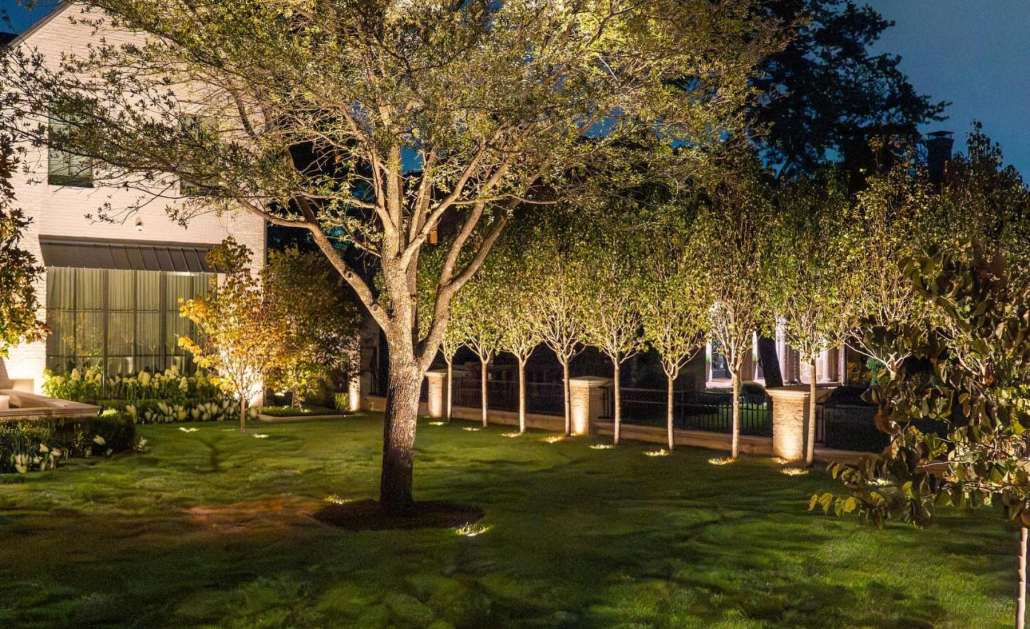 Elegant landscape lighting installation illuminates a lush green lawn, highlighting trees, shrubs, and a brick fence. The soft, warm light creates a welcoming and sophisticated ambiance around the property.