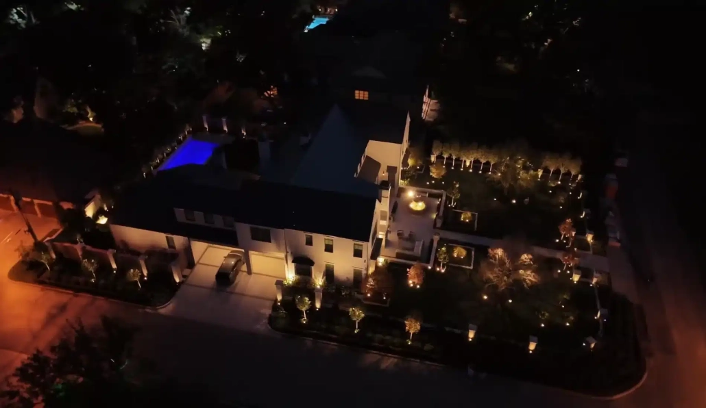Aerial view of a luxurious home at night, showcasing elegant outdoor lighting that illuminates the manicured garden and pool area, enhancing the home's curb appeal.