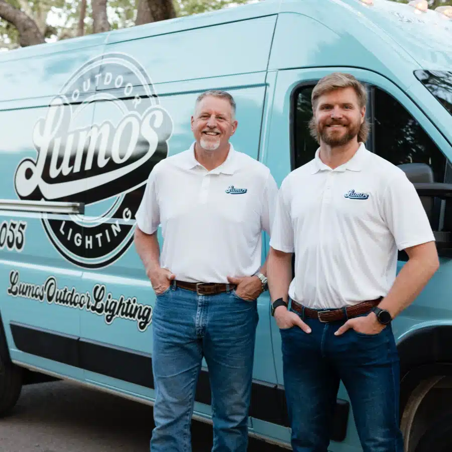 Two men from Lumos Outdoor Lighting stand proudly in front of their company van. The light blue van features the Lumos logo. Both men wear white polo shirts and jeans, smiling confidently.