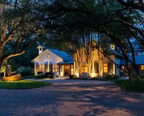 Evening view of a warmly lit ranch-style house with shuttered windows, large trees arching over a gravel driveway and a lit bear sculpture on a log.