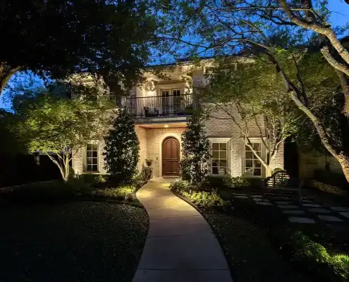 Two-story brick house at dusk with warm exterior lights, curved path leading to an arched wooden front door, trees and landscaped yard.