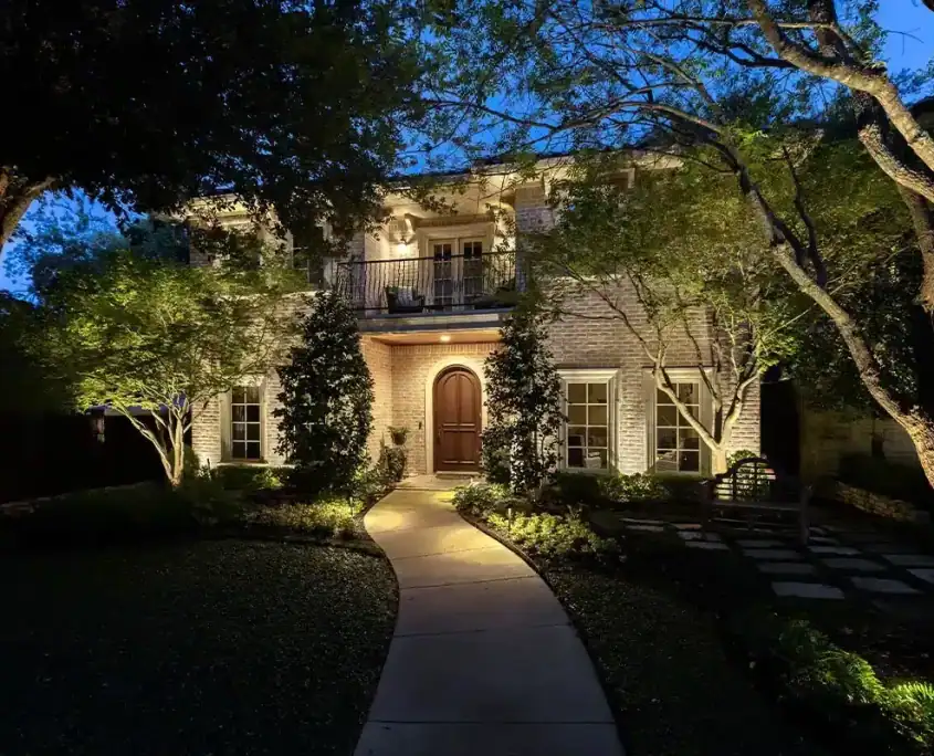 Two-story brick house at dusk with warm exterior lights, curved path leading to an arched wooden front door, trees and landscaped yard.