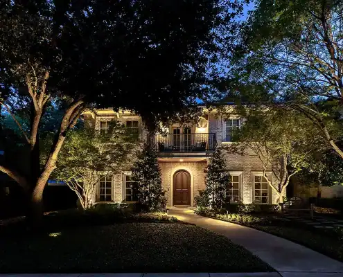 Two-story brick house at dusk with warm exterior lights, arched front door, second-floor balcony and trees framing a lit curved pathway.