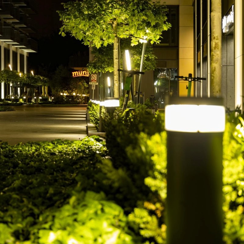 Nighttime commercial landscape lighting illuminates a pedestrian walkway. Modern light fixtures glow amidst lush greenery, enhancing the urban environment. A "muni" sign is visible in the background.