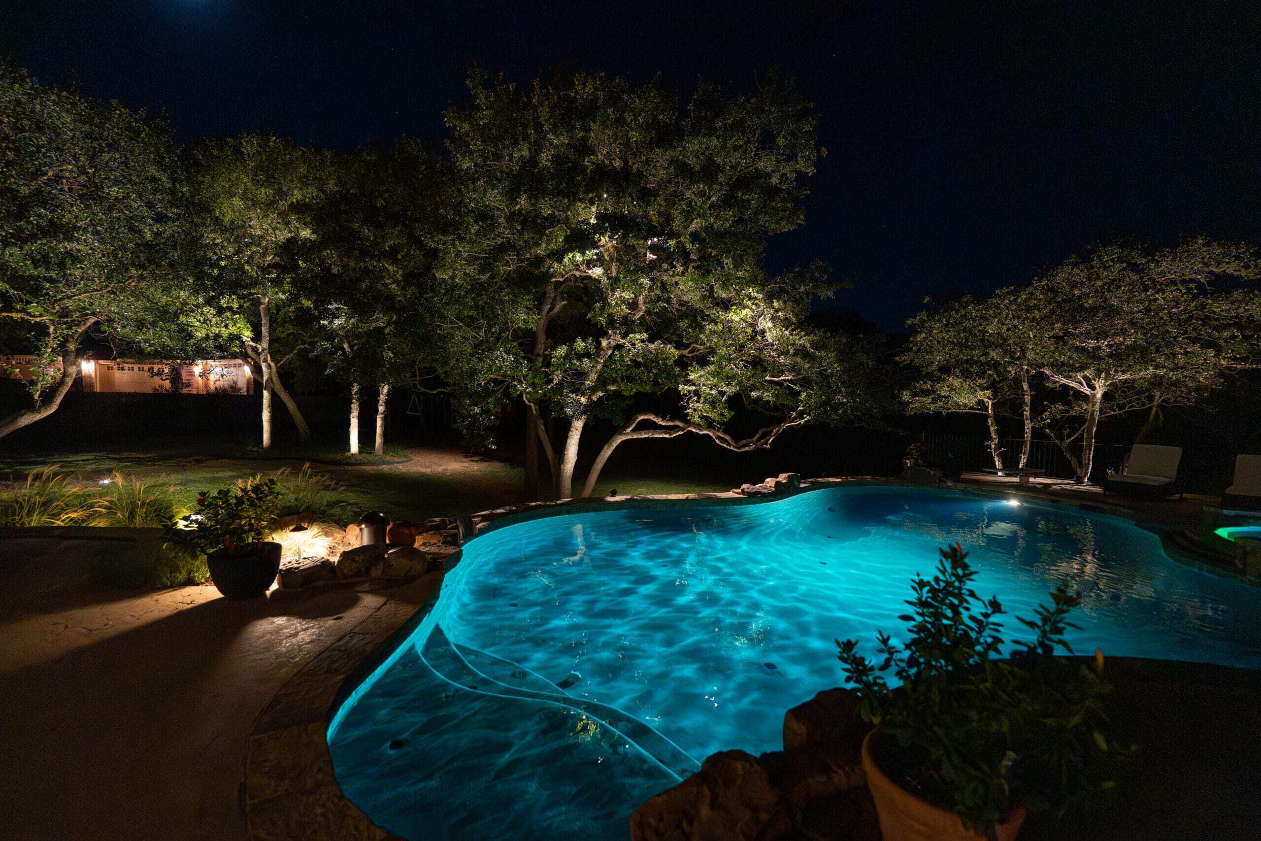 Outdoor lighting design illuminates a backyard pool at night. Trees surrounding the pool are lit from below, casting an inviting glow over the water and landscape.