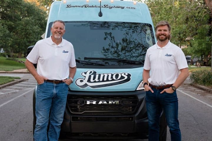 Two men stand proudly in front of a Lumos Outdoor Lighting van, promoting outdoor lighting maintenance services. The light blue van features the company logo and website.