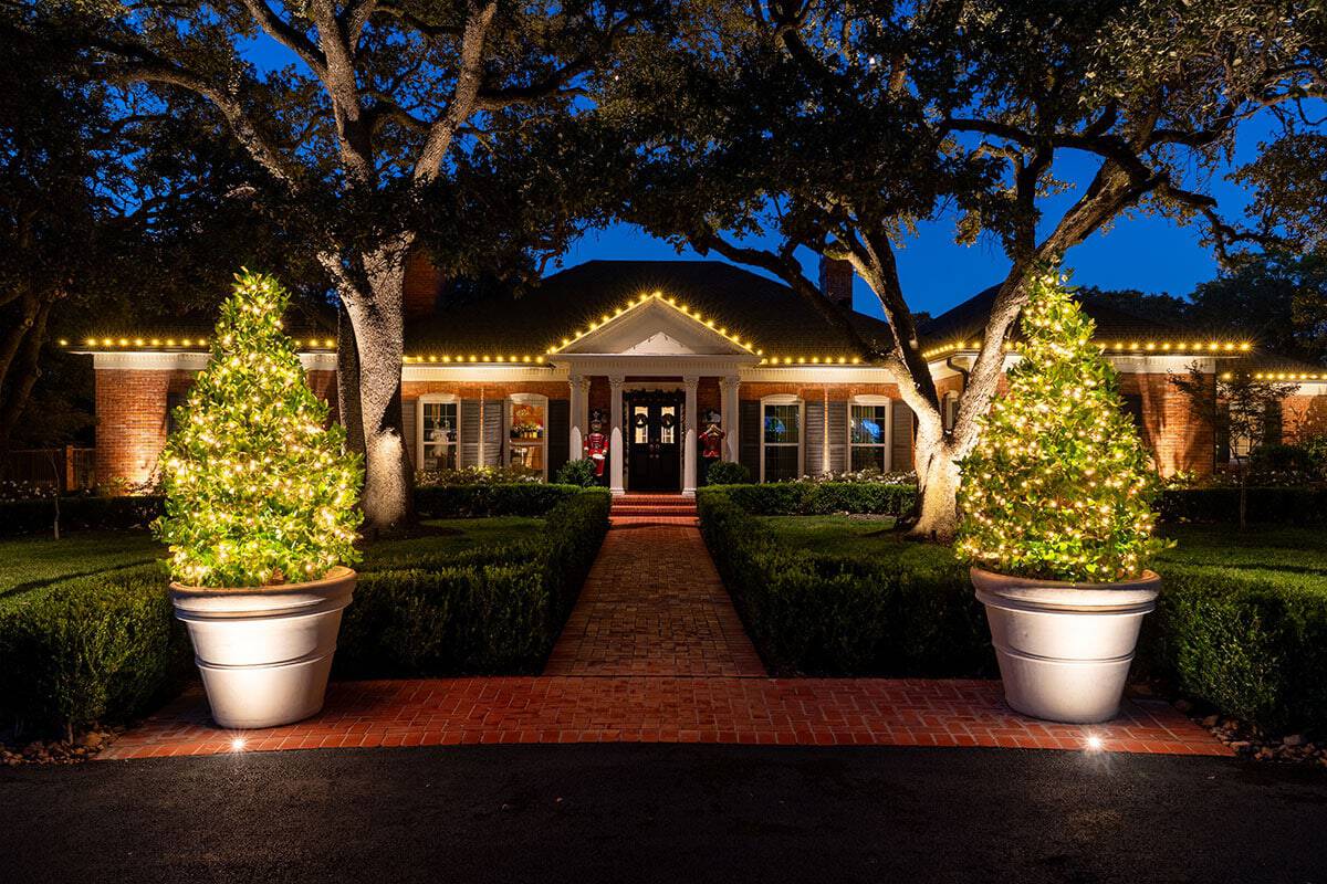Ridgeview home exterior at night, decorated for Christmas with lights outlining the roofline and two large potted Christmas trees flanking the brick walkway.