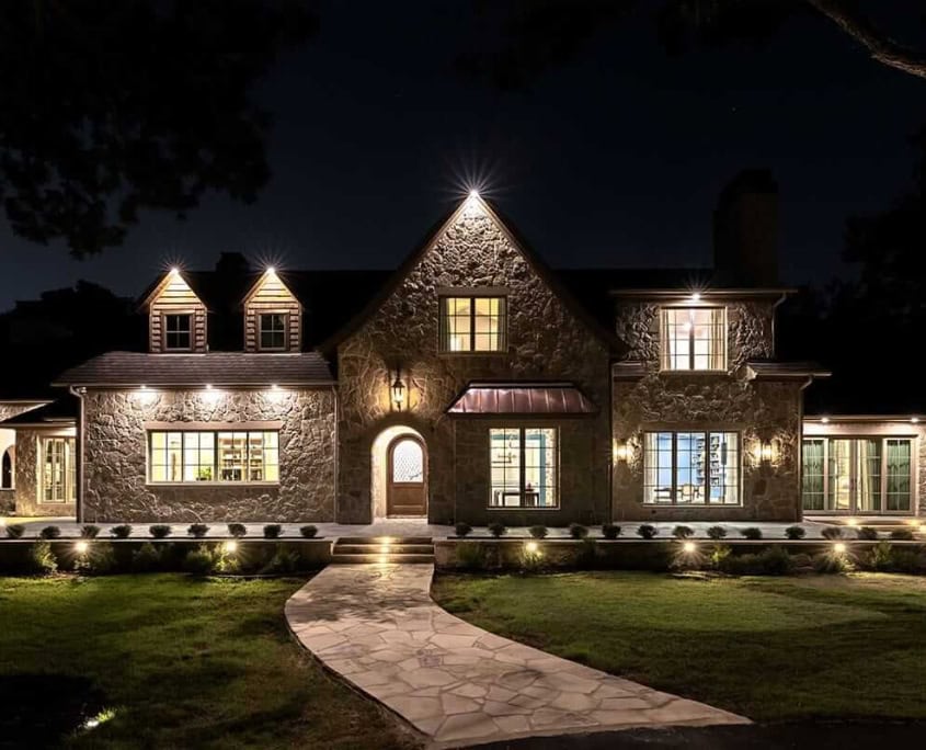 Large stone house at night, exterior lights and warm windows, curved stone walkway leading to arched front door and lawn.