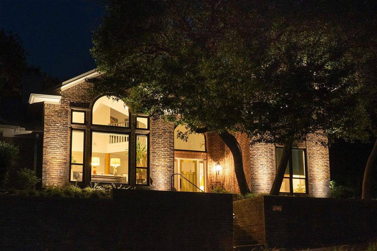 Elegant brick home at night, part of The Woodbriar Project, with illuminated arched windows and lush tree in the front yard. Warm light spills from the interior, creating a welcoming ambiance.