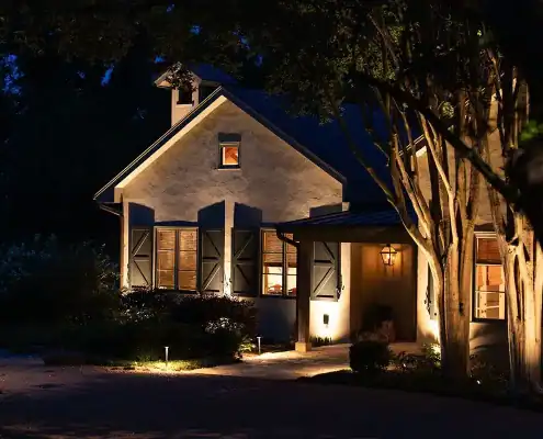Nighttime exterior of a stucco house with warm interior lights, closed shutters, a lit porch lantern, glowing pathway lights and nearby trees.