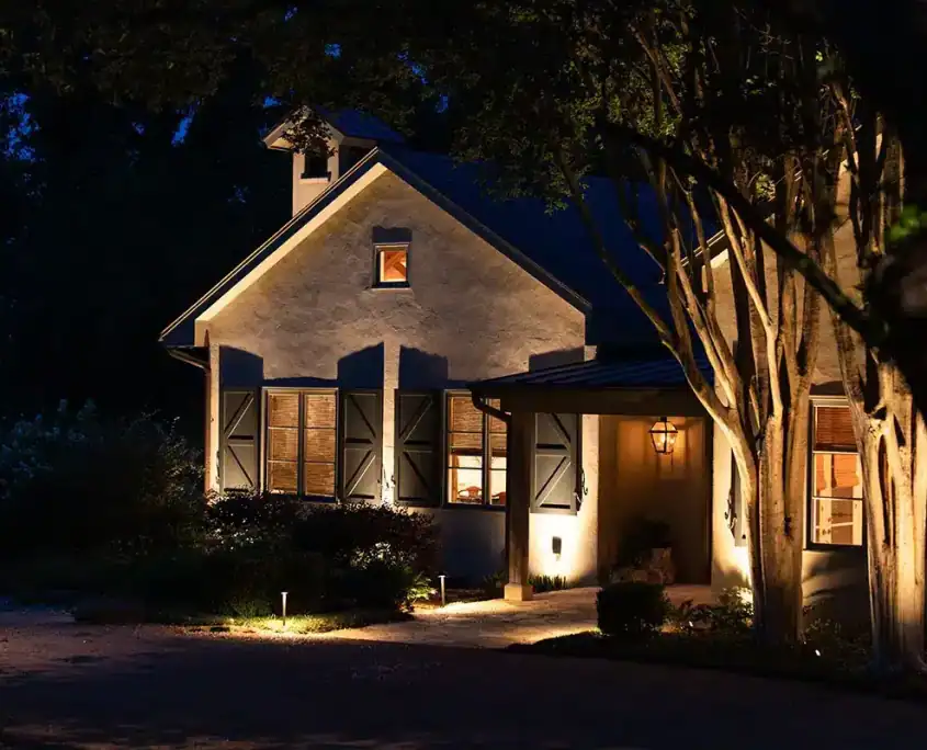 Nighttime exterior of a stucco house with warm interior lights, closed shutters, a lit porch lantern, glowing pathway lights and nearby trees.