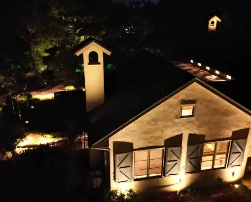 Night aerial view of a stucco house with lit windows, shuttered facade, small cupola tower, and illuminated garden paths.