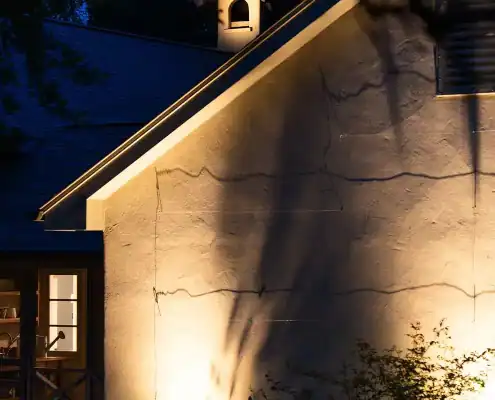 Night view of a stucco house gable lit by warm lights casting tall tree shadows; a lit window reveals a kitchen sink and shelves.