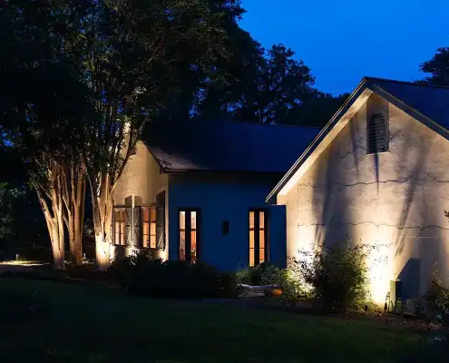 Night view of a lit stucco house with warm interior lights, uplighting on trees and exterior walls, and a deep blue evening sky.