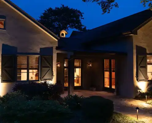 Night view of a lit house exterior with shuttered windows, warm indoor lights, wall lanterns, a stone walkway and garden landscape.