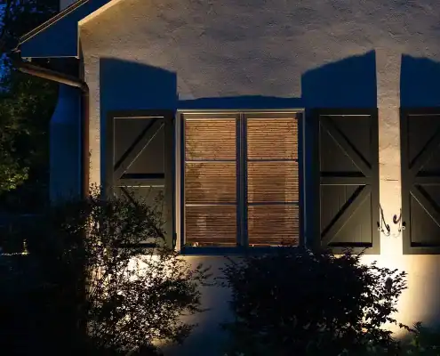Night exterior of a house: three closed dark shutters, center window glowing behind blinds, warm uplighting on stucco and front bushes.