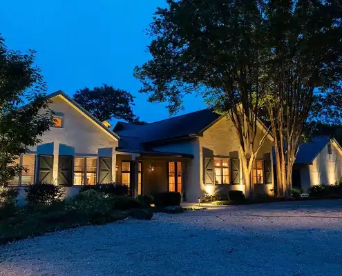 One-story house at dusk with warm interior lights, shutters, lit trees and shrubs, and a gravel driveway