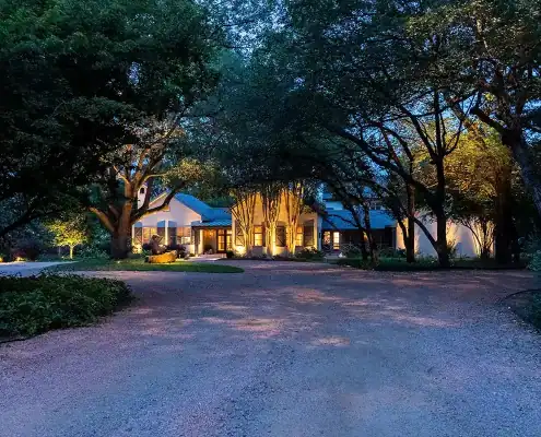 Large house at dusk with warm exterior lights, framed by tall trees and a wide gravel circular driveway.