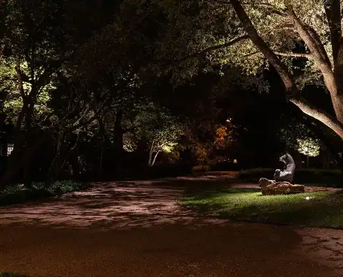 Nighttime curved driveway lined with trees, spotlight illuminating a seated animal statue on a rock beneath a large tree.