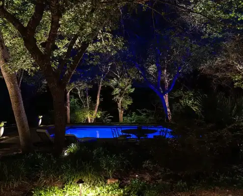 Nighttime backyard with a glowing blue swimming pool, patio chairs and table, and trees illuminated by ground lights.