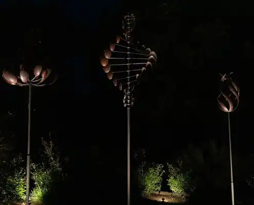 Three tall metal wind sculptures on poles at night, each with distinct petal-like shapes, spotlighted from below against a dark background.