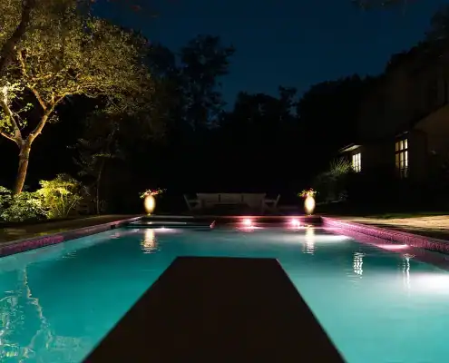 Nighttime backyard swimming pool lit from underwater, diving board in foreground, illuminated tree, glowing planters and a house with lit windows.