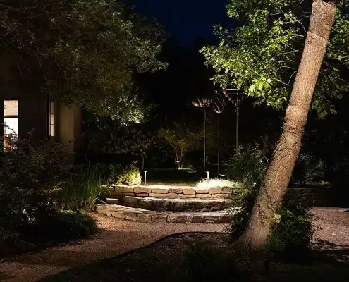Night garden scene with gravel path and tiered stone steps lit by landscape lights, trees and a glowing house window in background.