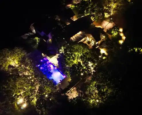 Aerial night view of a house with a blue-lit swimming pool surrounded by trees and warm pathway lights.