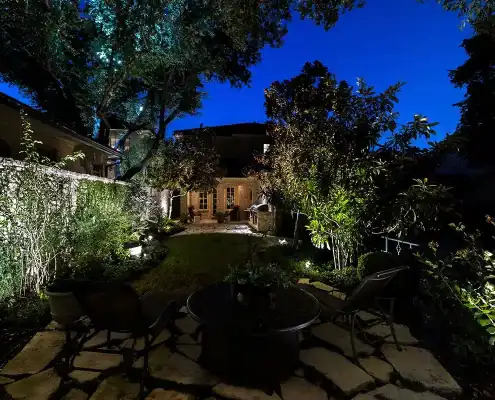 Night backyard patio with stone pavers, round table and two chairs, illuminated trees and shrubs, pathway to a warmly lit house under deep blue sky.