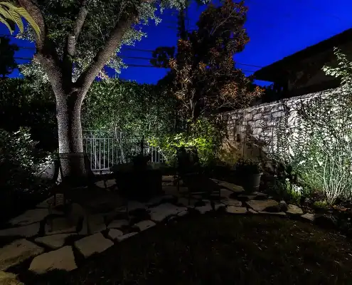 Nighttime backyard patio with illuminated tree, flagstone path, metal chairs, potted plants and vine-covered stone wall under deep blue sky.