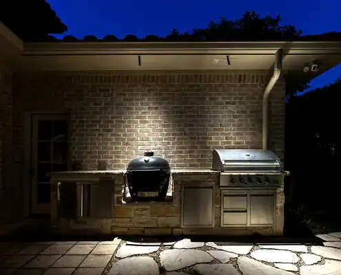 Outdoor stone and stainless-steel built-in grill island with two grills under a spotlighted brick wall at night.