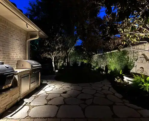 Nighttime backyard patio with irregular stone pavers, built-in grill on left, illuminated trees, plants and stone wall fountain on right.