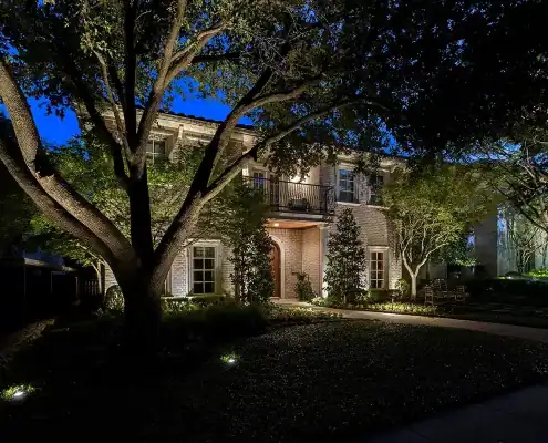 Two-story brick house at night with illuminated front entry and balcony, large tree in foreground and pathway lit by landscape lights.