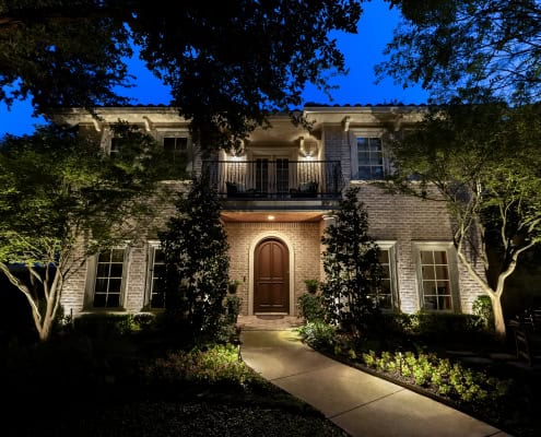 Night view of a two-story brick house entrance with arched wooden door, lit porch, balcony and trees framing the facade.