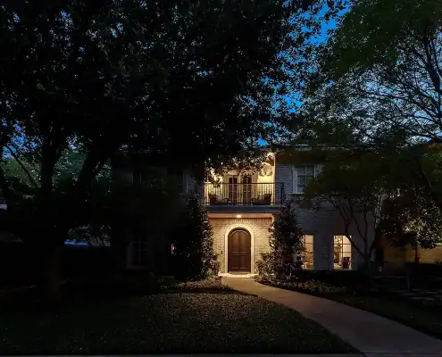 Two-story brick house at dusk, warmly lit arched front door and upstairs balcony, concrete path leading through shadowed trees and lawn.