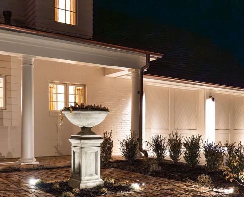 The Clover Ridge Outdoor Lighting Project Exterior view of Clover Ridge at night, showcasing elegant white brick, illuminated landscaping, and a classic stone planter. Warm light glows from the windows, highlighting architectural details.