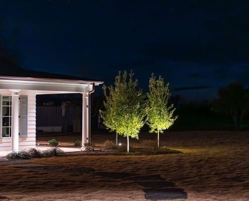 The Clover Ridge Outdoor Lighting Project Night view of Clover Ridge home exterior with illuminated trees. Warm light highlights the white siding, porch, and manicured lawn, creating a welcoming ambiance.