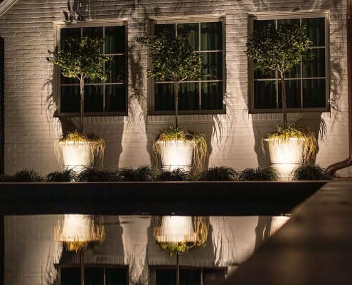 Exterior of Clover Ridge home at night. Three potted topiary trees are illuminated against a white brick wall, reflected in a pool. Modern and elegant outdoor design.